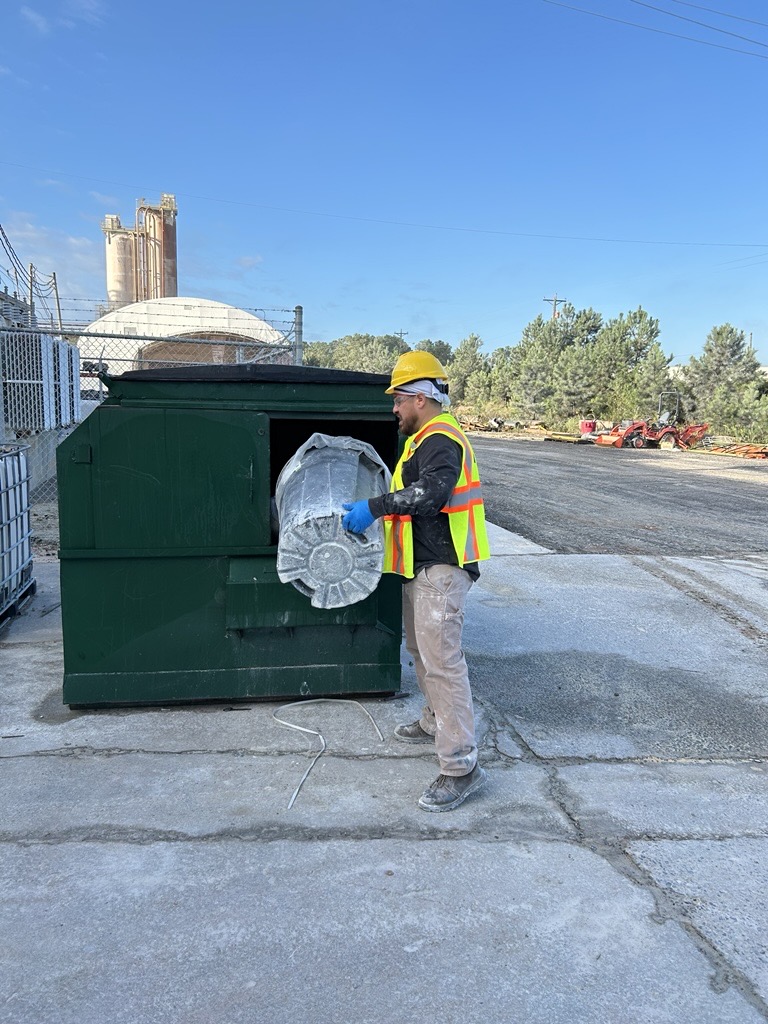 A worker wearing a hard hat and high-visibility vest places a large wrapped item into a green dumpster at an outdoor industrial facility.