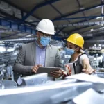 Two professionals wearing hard hats and face masks review information on a tablet while standing among metal equipment in a factory setting.