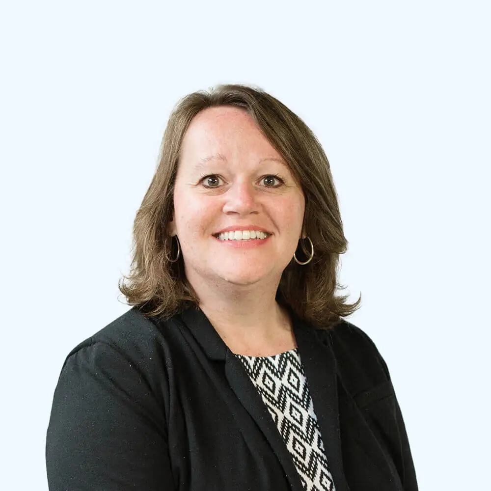 Portrait of a smiling woman with shoulder-length brown hair, wearing a black blazer over a patterned top, against a clean, light background.
