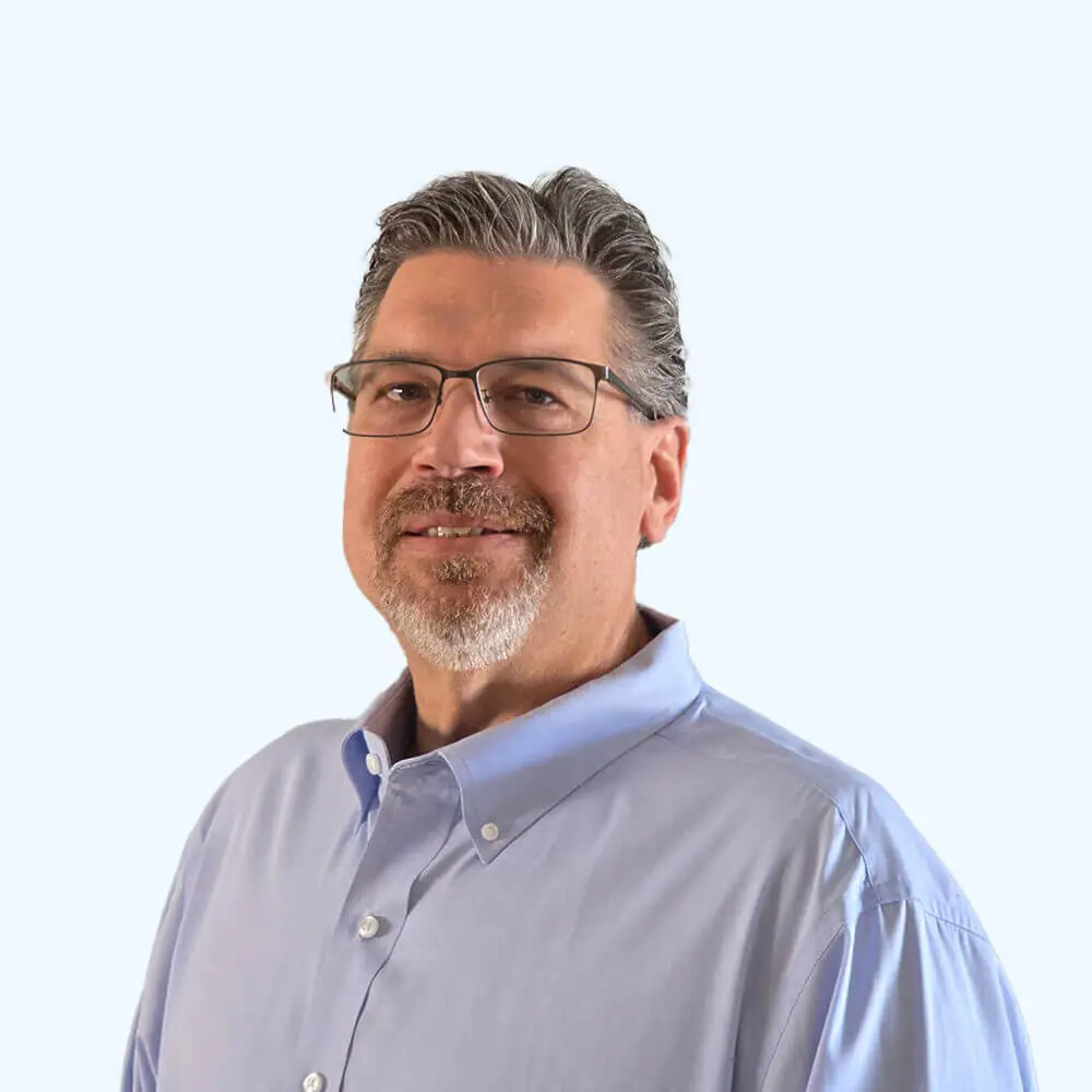 Portrait of a man with glasses, salt-and-pepper hair, and a short goatee, wearing a light blue button-down shirt against a clean, light background.