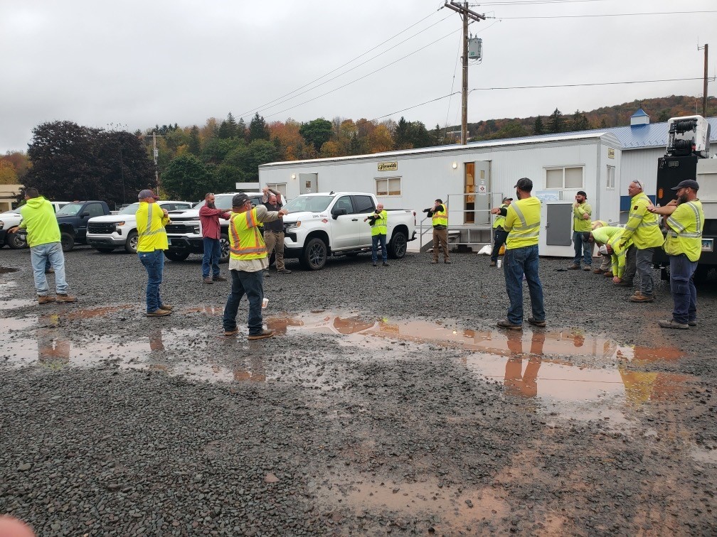 Group of workers in safety vests stretching together outdoors near trucks and building
