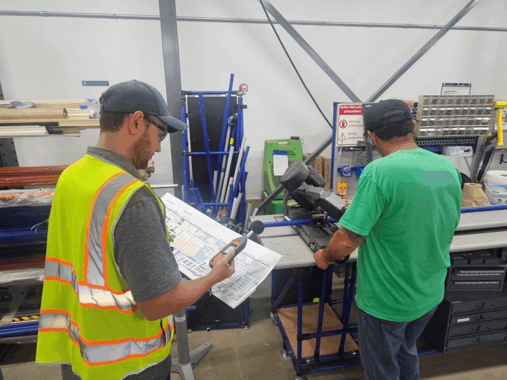 A worker in a high-visibility vest uses a sound level meter and checklist while another operates machinery in a workshop, demonstrating workplace noise monitoring.