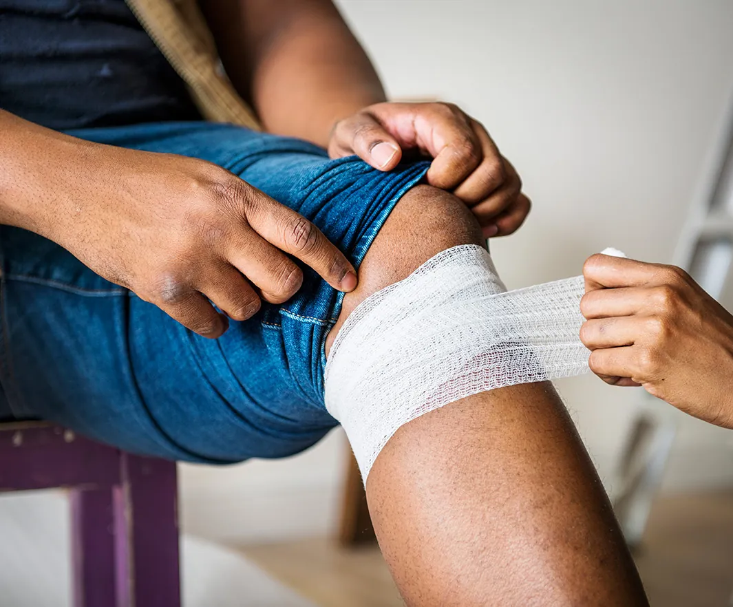 Close-up of a person sitting while another wraps a white gauze bandage around their injured knee, showing care and first aid attention.