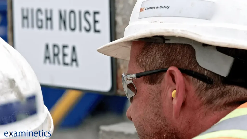 A close-up side view of a white male construction worker wearing a white hard hat with a sticker that reads ‘Leaders in Safety’. He is also wearing clear safety glasses and a yellow earplug in his right ear. His head is turned to the right, and the left side of his face is visible. Another worker's white hard hat is partially visible on the left. In the background, a white sign with a black frame reads ‘HIGH NOISE AREA’ in large text, which is slightly out of focus. A blue ‘examinetics’ logo is in the bottom left corner.