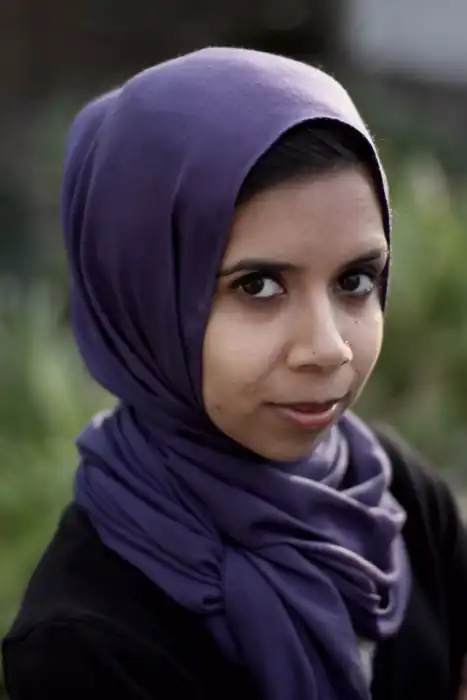 A portrait of a young woman with dark eyes, looking at the camera with a slight smile. She is wearing a purple hijab that frames her face and a black shirt. The background is blurred and green, suggesting she is outdoors or in a garden.
