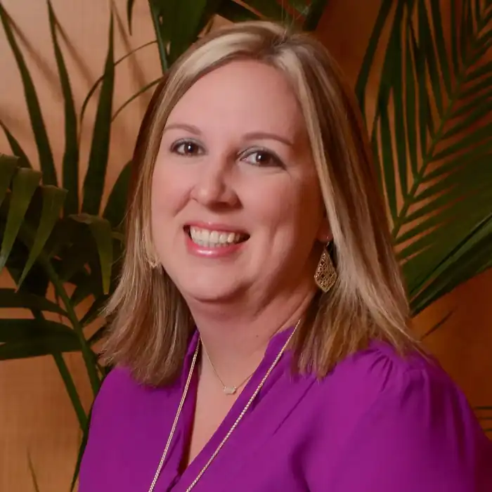 A professional headshot of a smiling white woman with blonde hair, wearing a vibrant purple blouse. She has a necklace with a small pendant and large, gold, filigree earrings. She is looking directly at the camera with a wide smile. The background is a mix of a brown wall and a large green plant with long leaves.