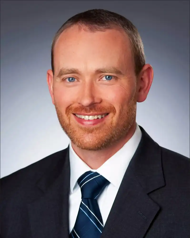 A professional headshot of a smiling white man with blue eyes, a short red beard, and a receding hairline. He is wearing a dark suit jacket, a white collared shirt, and a dark blue tie with white pinstripes. He is looking directly at the camera with a bright, friendly smile. The background is a gradient from dark grey to light grey.