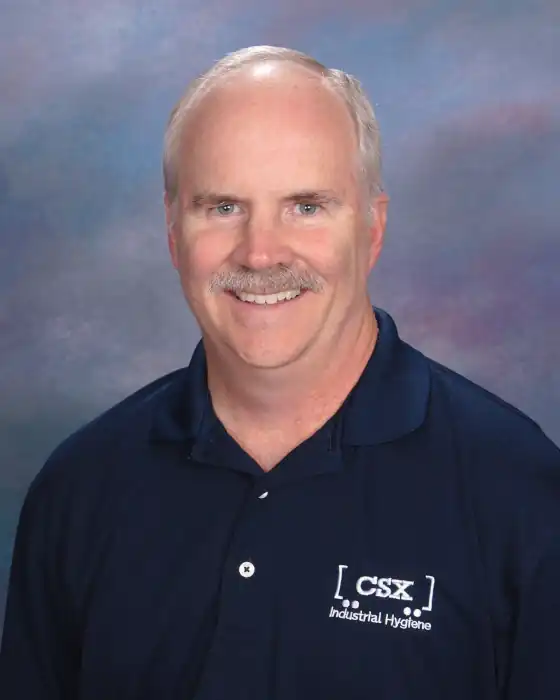 A professional headshot of a smiling white man with a bald head and a grey mustache. He is wearing a dark blue polo shirt with a white logo that reads ‘CSX Industrial Hygiene’. He is looking directly at the camera. The background is a multi-colored blend of blue, purple, and green.