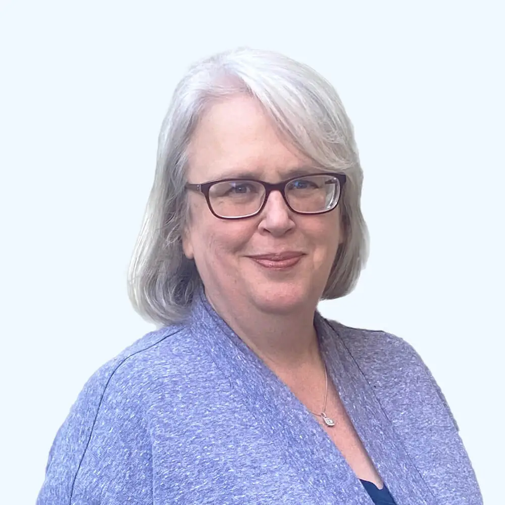 Portrait of a woman with shoulder-length gray hair and glasses, wearing a purple cardigan and necklace, smiling slightly against a clean, light background.