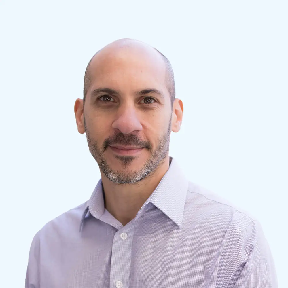 Portrait of a man with a shaved head and short beard wearing a light-colored button-down shirt, facing the camera against a plain, light background.