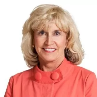 A professional headshot of a smiling white woman with blonde, shoulder-length hair and bangs. She is wearing a coral-colored jacket with large buttons on the collar. She has small earrings and is looking directly at the camera with a friendly smile. The background is a solid white.