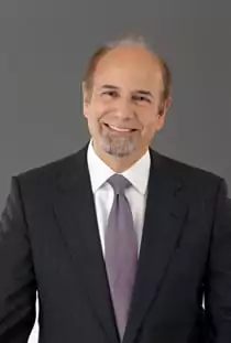 A professional headshot of a smiling white man with a short, grey goatee and a receding hairline. He is wearing a dark suit jacket, a white collared shirt, and a purple striped tie. He is looking directly at the camera. The background is a solid grey.