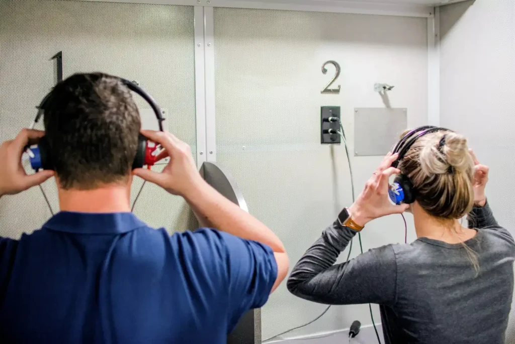 A person with short brown hair in a blue polo shirt and a person with blonde hair in a ponytail and a gray long-sleeve shirt are seen from behind, sitting in separate audiology booths. They are each holding headphones over their ears, preparing for a hearing test. The booths are numbered "1" and "2."