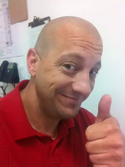 A smiling, bald man with a light-skinned complexion and a wide nose is giving a thumbs-up to the camera. He is wearing a red polo shirt and looking slightly down and to the left. The background is a plain white wall with a clipboard and a black phone visible behind him. The image appears to be a casual selfie.
