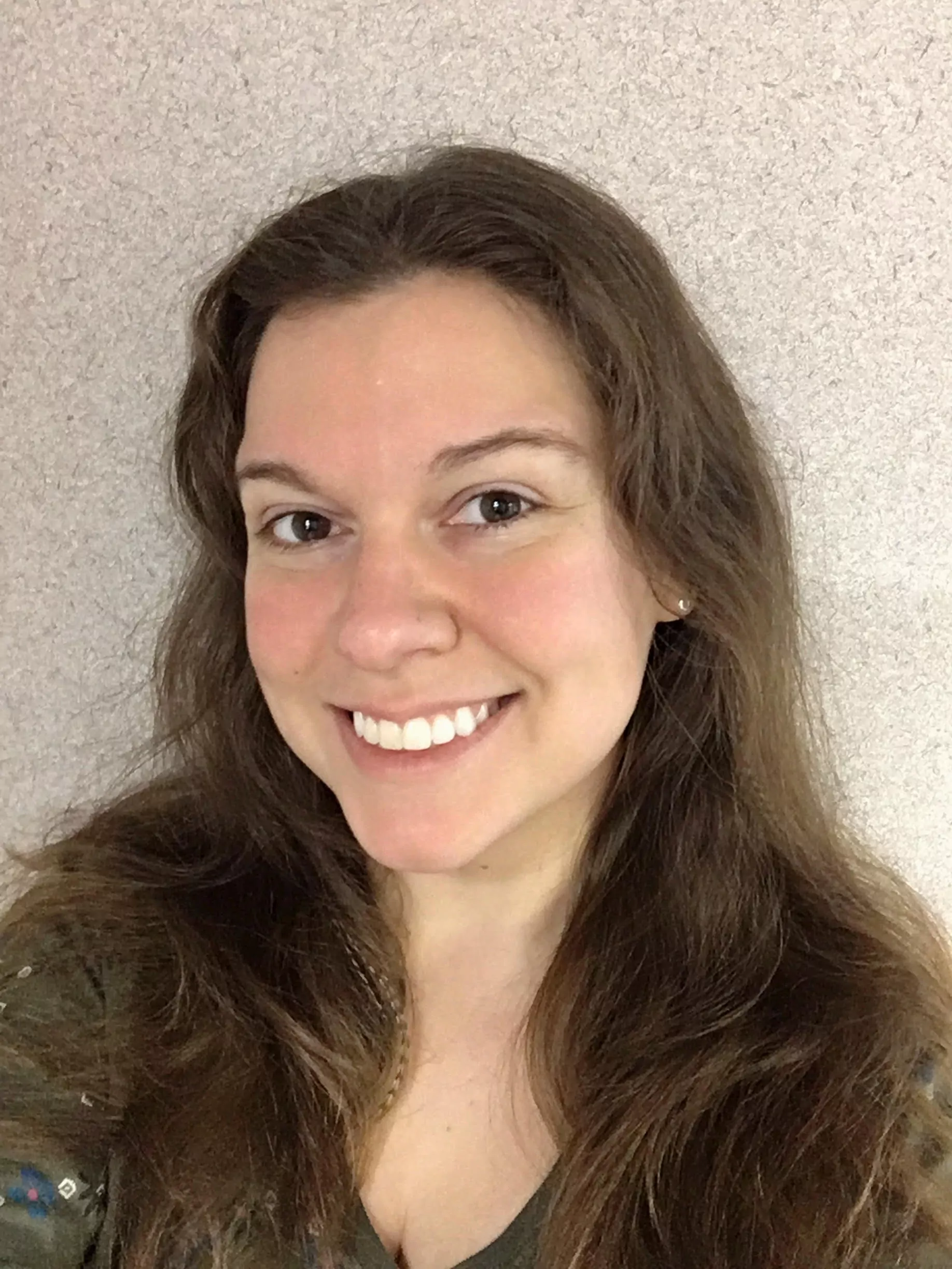A smiling white woman with long, brown, wavy hair and a slight part in the middle. She is wearing a green shirt and small stud earrings. She is looking directly at the camera with a bright, friendly smile. The background is a plain, light grey textured wall.