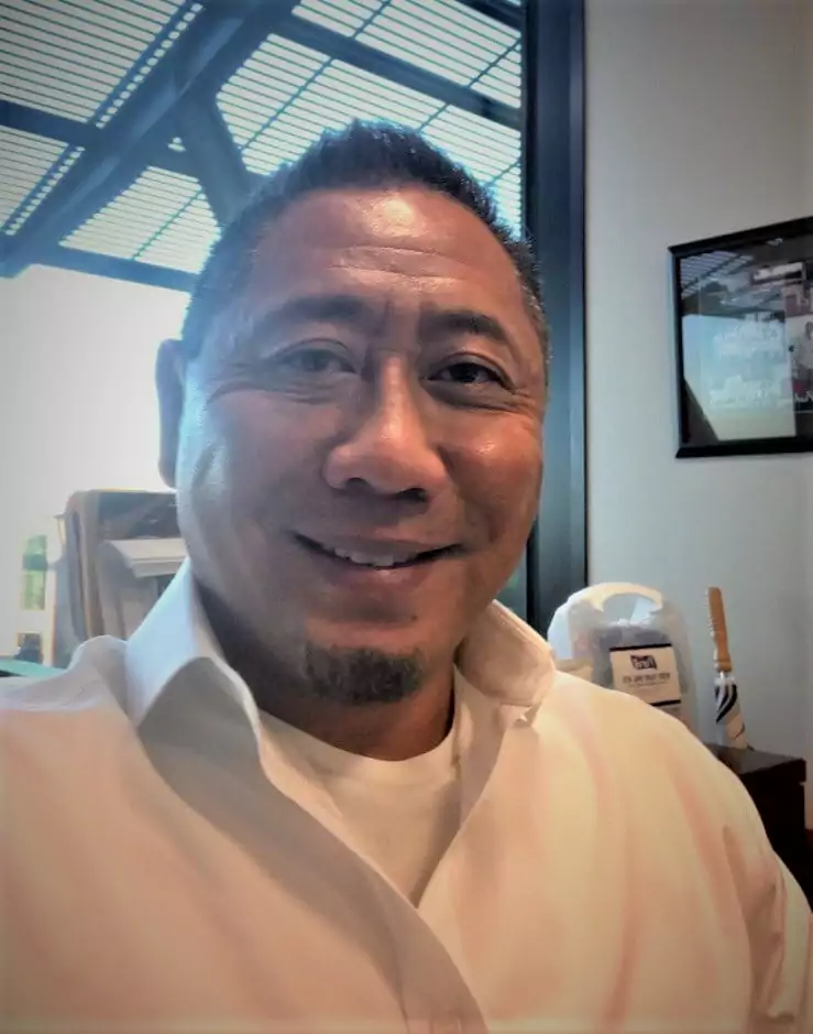 A selfie-style headshot of a smiling man of color with a neatly trimmed goatee and short, dark hair. He is wearing a white collared shirt. He is looking directly at the camera with a friendly expression. The background is an office setting, with a window and a framed picture on the wall.