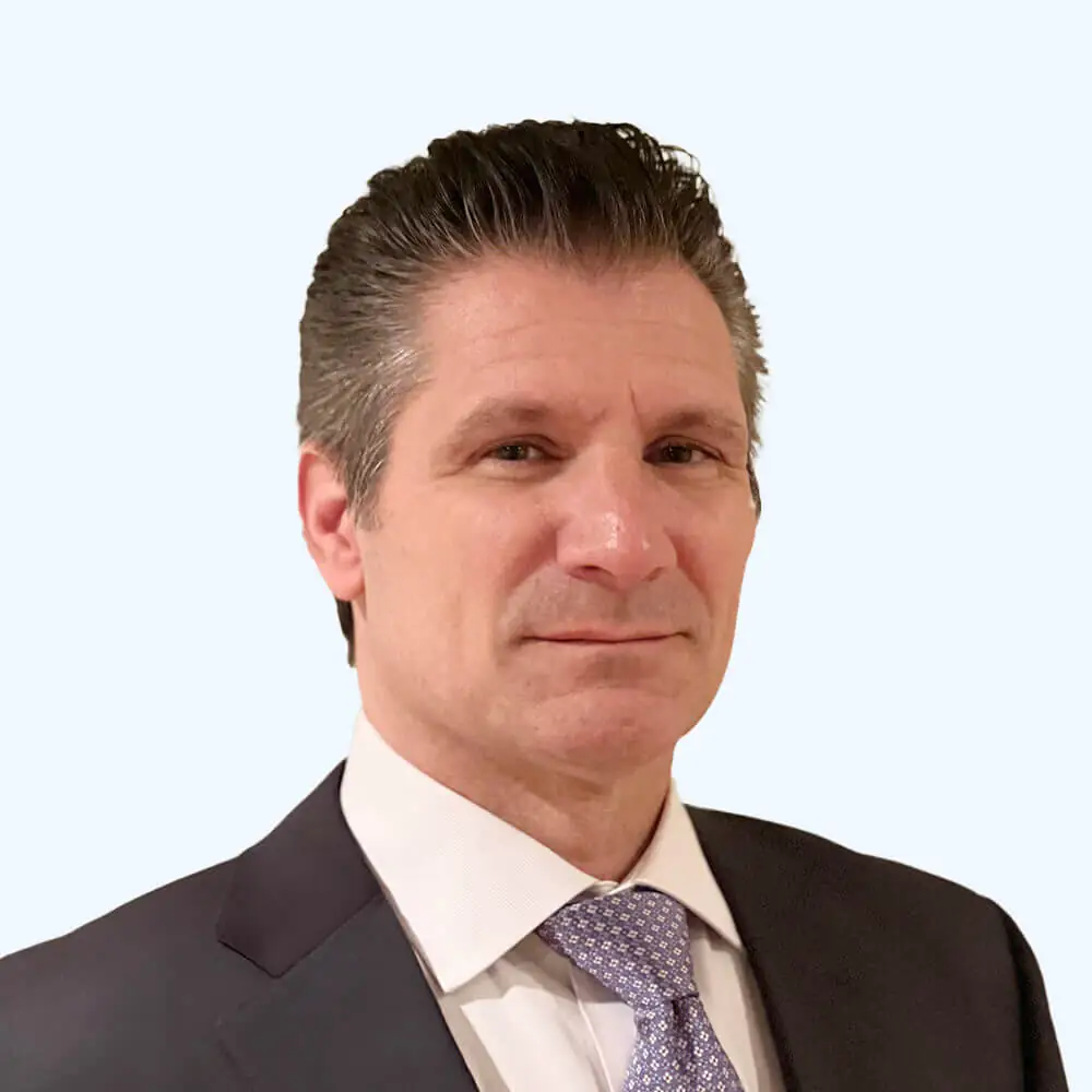 Portrait of a man with neatly styled dark hair, wearing a dark suit, white dress shirt, and patterned tie, facing slightly toward the camera against a clean, light background.