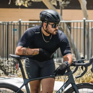 A portrait of a male cyclist with a beard and mustache, wearing a sleek black cycling outfit, sunglasses, and a black helmet. He is leaning on a black road bicycle, resting his forearms on the handlebars, and looking off to his right with a serious expression. He has a gold chain around his neck and a watch on his left wrist. His right arm has a large tattoo. The background is an urban setting with a metal fence and blurred trees.