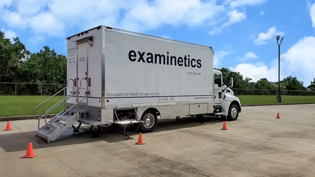 A large white Examinetics mobile medical unit truck is parked in a parking lot under a blue sky with white clouds. It has stairs leading up to a side door and is labeled with "examinetics" and "Occupational Health Screening Unit." Several orange traffic cones are visible around the truck.