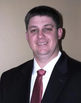 A professional headshot of a smiling white man with short, dark hair. He is wearing a black suit jacket over a white collared shirt and a dark red tie. He is looking directly at the camera with a friendly expression. The background is a plain, light brown wall.