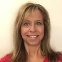A professional headshot of a smiling white woman with long, layered brown hair with blonde highlights and bangs. She is wearing a red top and is looking directly at the camera with a warm smile. The background is a plain, off-white wall.