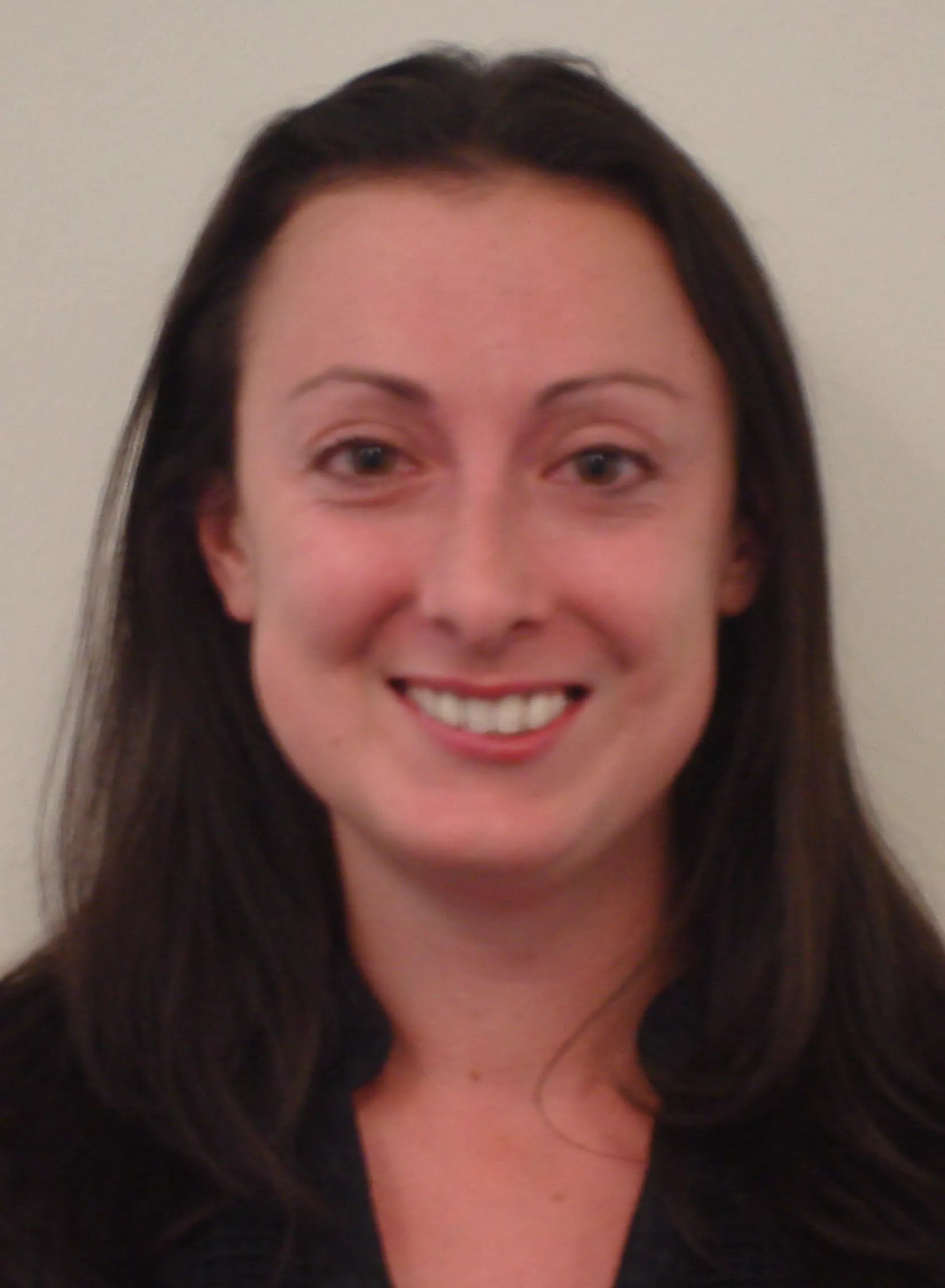 A professional headshot of a smiling white woman with long, dark brown hair parted on the side. She is wearing a dark top and looking directly at the camera with a bright smile. The background is a plain, light-colored wall.