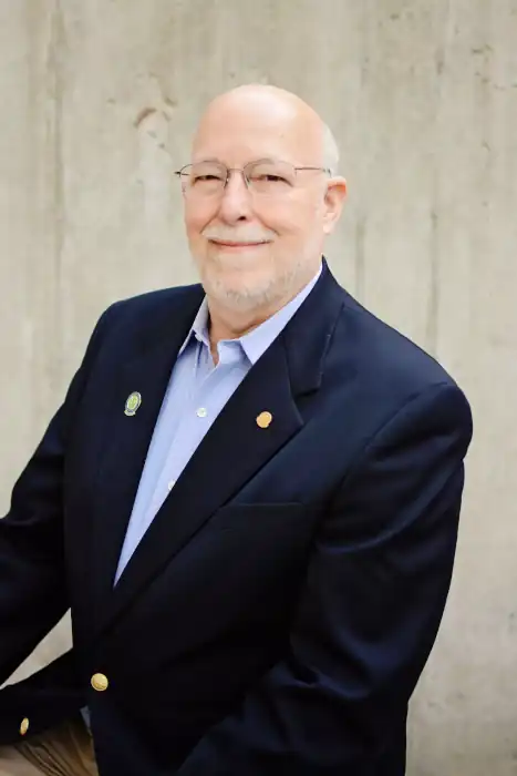 A portrait of an older white man with a white goatee, a bald head, and glasses. He is wearing a dark blue blazer with two small pins on the lapel, and a light blue collared shirt underneath. He is smiling warmly at the camera. The background is a plain, light grey concrete wall.