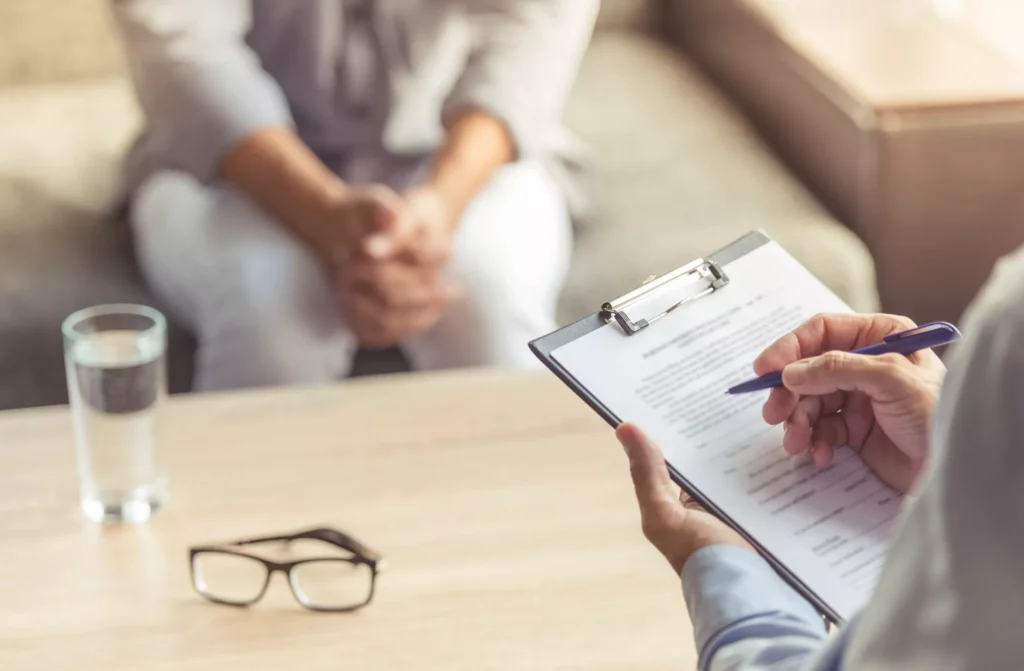 A medical professional, seen from the back, holds a clipboard and a pen, writing on a form. In the foreground on the table are a glass of water and a pair of eyeglasses. In the blurry background, a person is seated on a couch with their hands clasped.