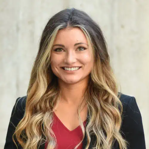 A portrait of a smiling white woman with long, wavy blonde and brown hair and light-colored eyes. She is wearing a black blazer over a red top and is looking directly at the camera with a wide smile. The background is a plain, light grey concrete wall.