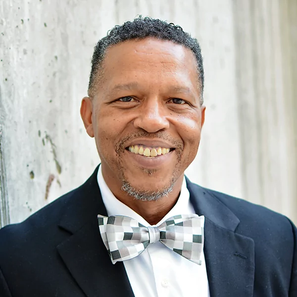 A smiling Black man with a salt-and-pepper beard and short, curly hair. He is wearing a black suit jacket, a white collared shirt, and a grey checkered bow tie. He is looking at the camera and smiling broadly. The background is a textured grey concrete wall.