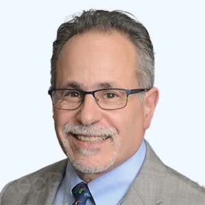 Portrait of a smiling man with glasses, gray hair, and a trimmed mustache and goatee, wearing a suit jacket, dress shirt, and tie against a light background.