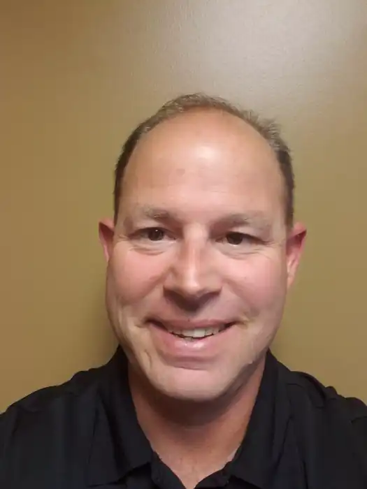 A headshot of a smiling white man with a receding hairline and short, brown hair. He is wearing a dark, possibly black, button-up shirt and is looking directly at the camera with a friendly smile. The background is a plain, light brown wall.