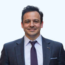 Portrait of a man with short dark hair and light stubble, wearing suit in front light blue backdrop