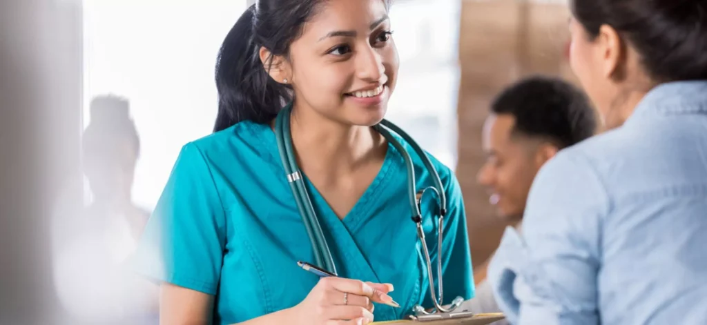 A Hispanic female medical professional, possibly a nurse or a physician's assistant, is smiling while looking at a patient. She has dark hair tied back in a ponytail and is wearing a turquoise scrub top and a stethoscope around her neck. She is holding a clipboard and a pen, appearing to be taking notes or explaining a procedure. The patient's back is visible in the foreground, and another person, a man of color, is blurred in the background, also smiling.