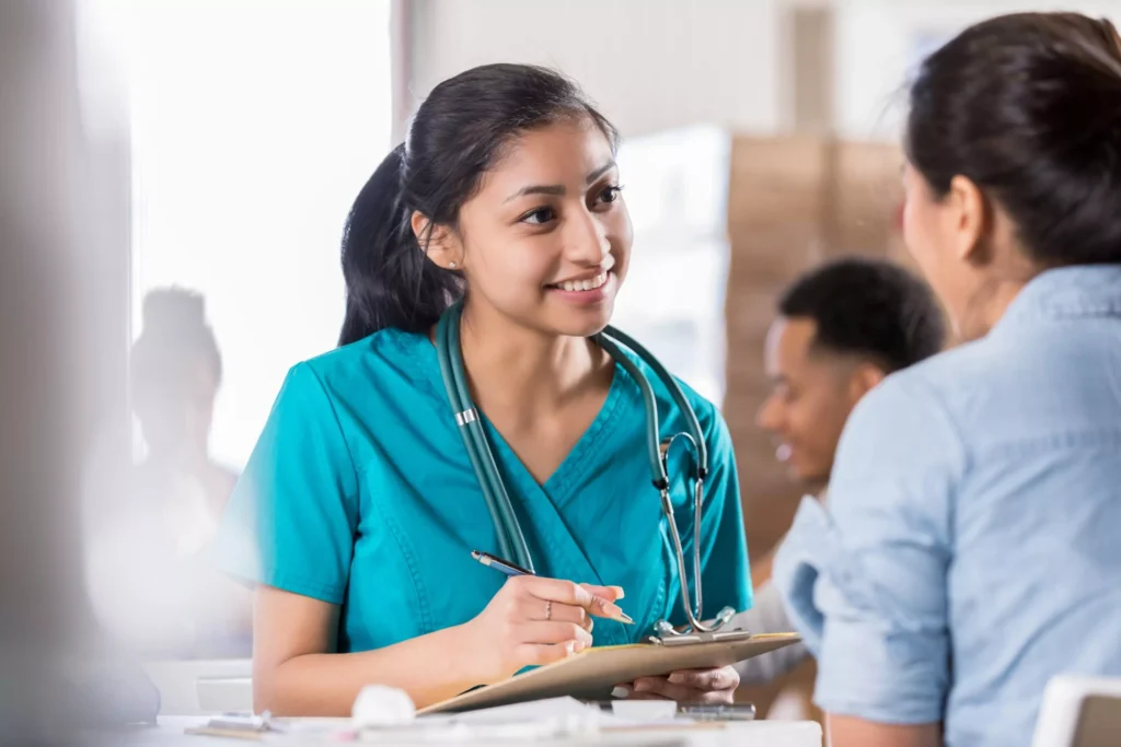 A young female healthcare professional wearing teal scrubs and a stethoscope around her neck smiles warmly while holding a clipboard and pen, engaging in conversation with a patient seated across from her in a casual clinical setting.
