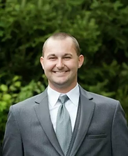 A professional headshot of a smiling white man with a short buzzcut and a small earring in his left ear. He is wearing a grey suit jacket, a matching vest, a white shirt, and a grey striped tie. He is looking directly at the camera with a wide smile. The background is a blurred green foliage.