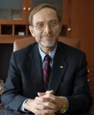 A portrait of an older white man with a small, neatly trimmed beard and mustache, and glasses. He is sitting at a desk with his hands clasped in front of him and is smiling. He is wearing a dark suit, a blue collared shirt, and a red striped tie. The background is a blurred office setting with a wooden bookshelf and an office chair.