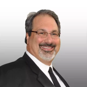 A man with graying hair and a neatly trimmed beard, wearing a black suit and tie, smiles broadly at the camera. He is wearing glasses.