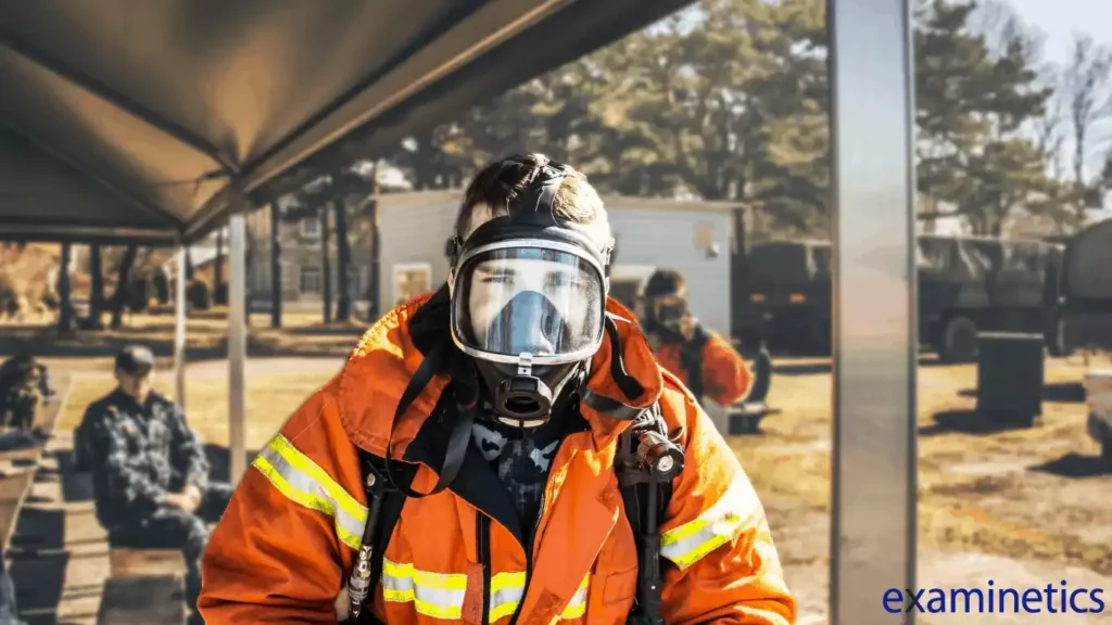 A person wearing a large, full-face respirator mask and a bright orange protective jacket with yellow reflective stripes. The person is looking directly at the camera with a determined expression. In the blurred background, other people in uniform are visible, along with military vehicles and trees. The scene appears to be an outdoor training or exercise. The bottom right corner has a blue ‘examinetics’ logo.