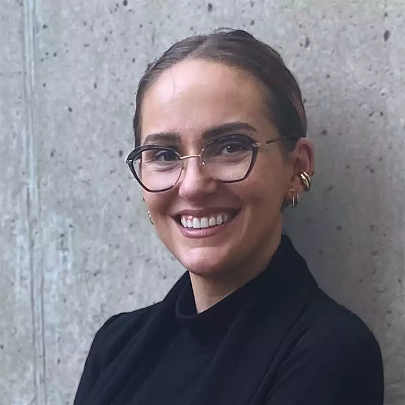 A close-up professional headshot of a smiling woman with dark brown hair pulled back. She is wearing stylish glasses with a black frame on top and a gold wire on the bottom. She also has multiple gold earrings. She is wearing a black turtleneck and a black blazer. She is looking at the camera with a wide smile. The background is a plain, light grey concrete wall.