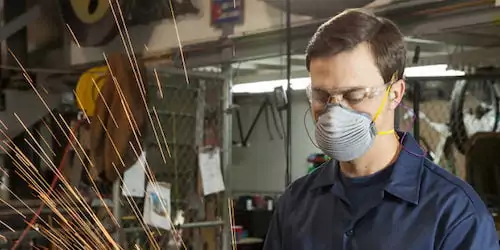 A male worker, who appears to be in his 30s, is wearing a pleated respirator mask, clear safety glasses, and yellow earplugs. He is wearing a dark blue work shirt and is looking down at his work. Sparks are flying toward the left side of the frame, indicating that he is welding or grinding metal. The background is a cluttered workshop with shelves, equipment, and a yellow hard hat visible.