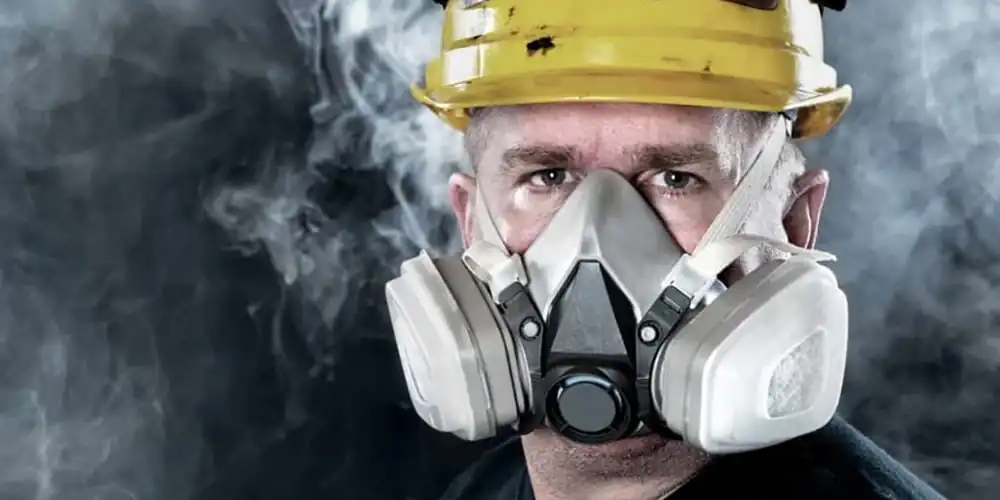 A close-up of a white male worker wearing a half-face respirator mask and a yellow hard hat. He is looking directly at the camera with a serious and intense expression. The background is dark and filled with a cloud of grey smoke or dust, emphasizing the need for the protective gear. The respirator mask has two large grey filters on either side and a valve in the center.