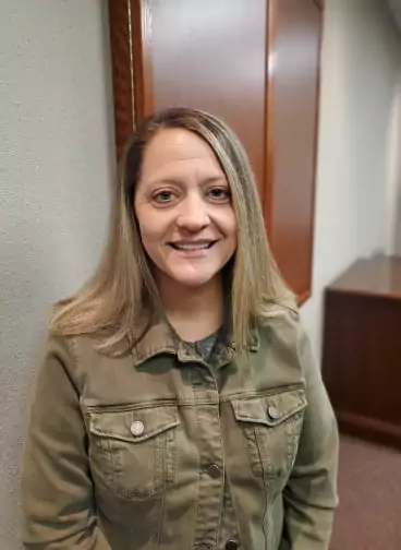 A woman with a warm smile, wearing a denim-style, olive green jacket. She has long, blonde hair that is parted in the middle. She is looking directly at the camera. The background is an indoor setting with light-colored walls and a dark wooden cabinet or door. The image appears to be a casual portrait or a selfie.