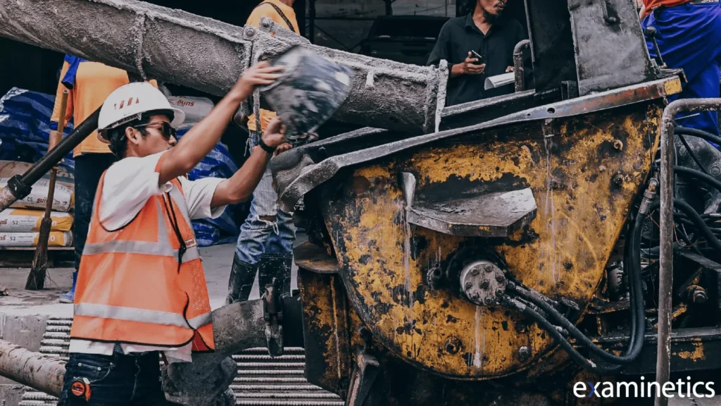 Construction worker in a white hard hat, orange safety vest, and sunglasses pouring a bucket of wet concrete into a large yellow industrial cement mixer, with other workers and construction materials visible in the background.