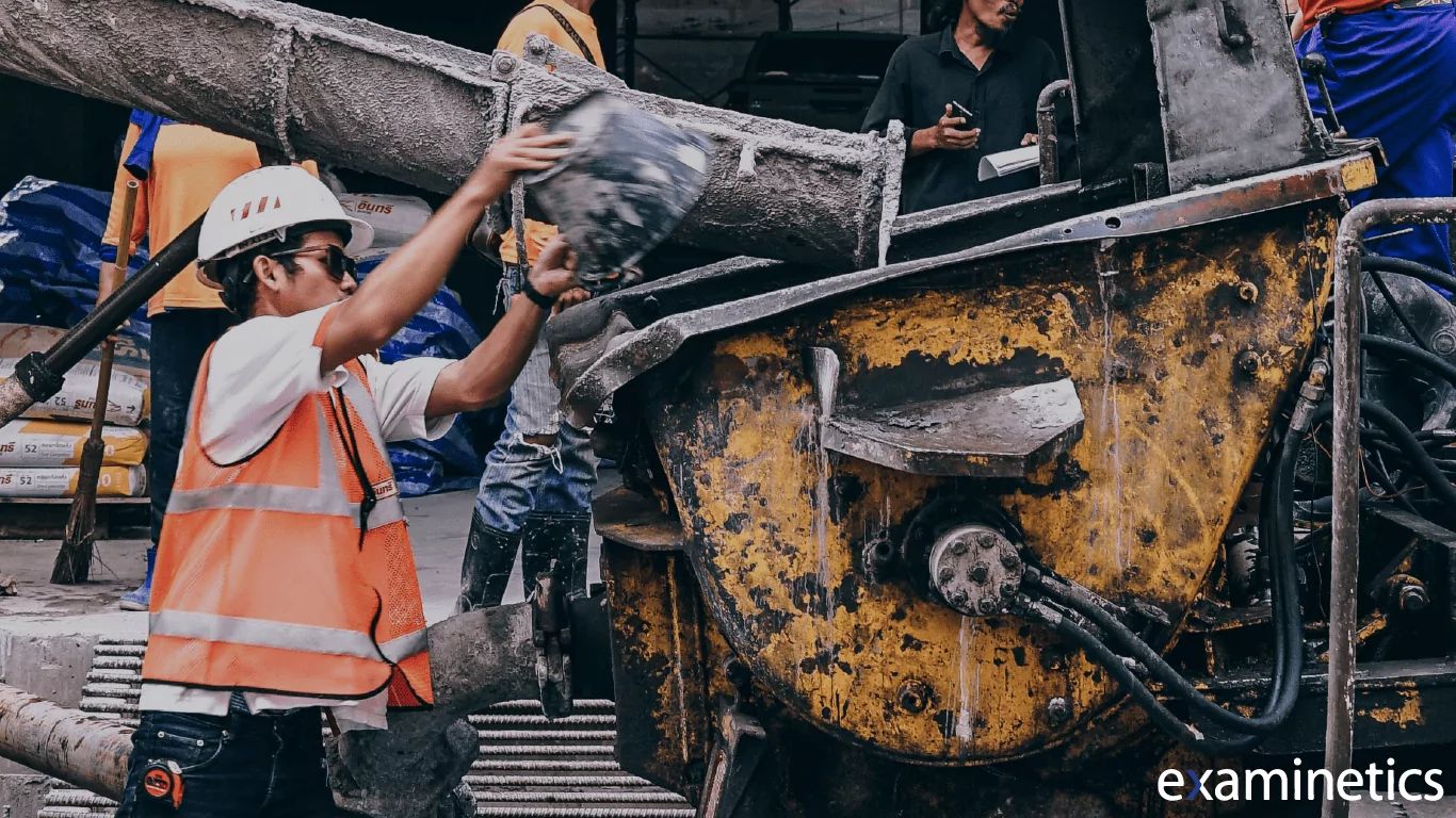 Construction worker in a white hard hat, orange safety vest, and sunglasses pouring a bucket of wet concrete into a large yellow industrial cement mixer, with other workers and construction materials visible in the background.