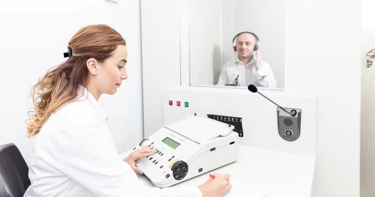 A female audiologist with light brown hair in a white lab coat is sitting at a desk, facing a soundproof booth. She is writing on a notepad while monitoring a patient. In the booth, a male patient wearing headphones is looking to the side, with his hand on the right ear of the headphones. The booth has a window, allowing the audiologist to observe the patient. A microphone is positioned between them, and the audiologist has a control panel and audiometer on the desk in front of her. The setting is clean and brightly lit.