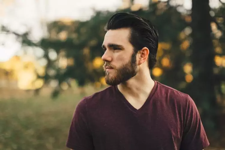 A young white man with a well-groomed, dark brown beard and slicked-back dark hair. He is wearing a simple maroon V-neck t-shirt. He is looking off to the left side of the frame with a thoughtful or serious expression. The background is a soft-focus outdoor setting with autumn foliage and trees.