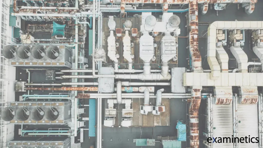 A top-down aerial view of an industrial rooftop filled with HVAC systems, large ventilation fans, ductwork, and interconnected pipes. The metal surfaces show visible rust and wear, with some pipes wrapped in insulation and others exposed. The layout is dense and organized, with rectangular and cylindrical components in varying sizes.