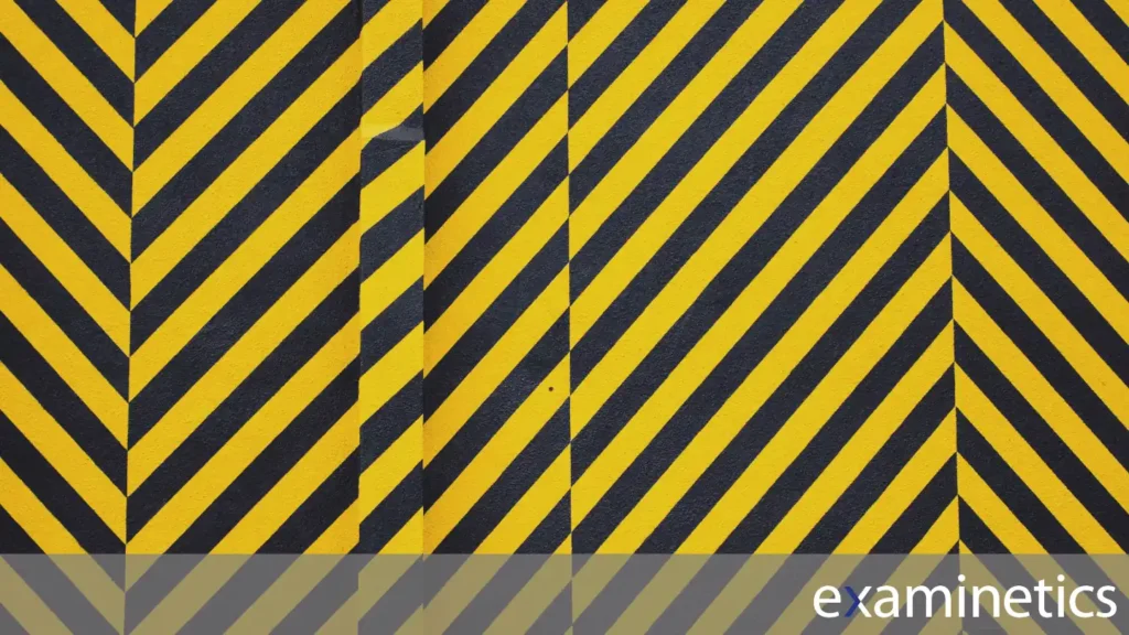 A full-frame shot of a wall painted with diagonal yellow and black warning stripes, which are commonly used to signify hazardous areas. The stripes are all angled from the top-right to the bottom-left. A vertical seam or column is visible in the middle of the wall, interrupting the pattern. The bottom of the image has a semi-transparent grey bar overlay with the white ‘examinetics’ logo in the bottom right corner.