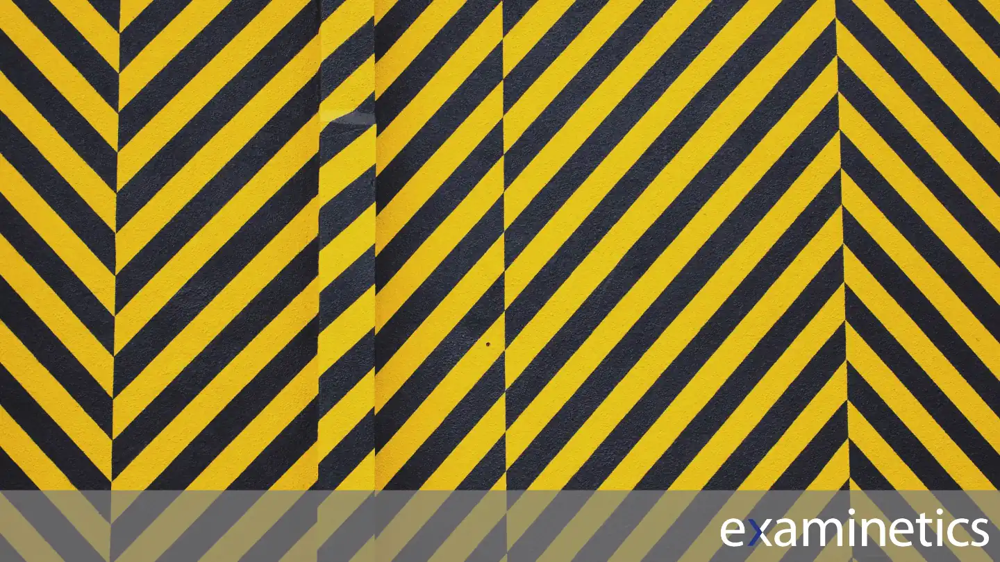 A full-frame shot of a wall painted with diagonal yellow and black warning stripes, which are commonly used to signify hazardous areas. The stripes are all angled from the top-right to the bottom-left. A vertical seam or column is visible in the middle of the wall, interrupting the pattern. The bottom of the image has a semi-transparent grey bar overlay with the white ‘examinetics’ logo in the bottom right corner.